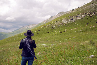 Woman shepherd standing in the mountains in a meadow in a hat with sheep