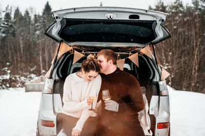 Young couple holding wineglasses standing by car during winter