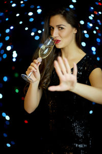 Portrait of beautiful young woman drinking champagne while gesturing against illuminated lights