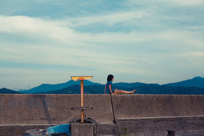 Side view of boy sitting on retaining wall against mountains