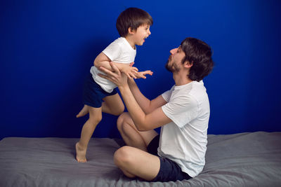 Boy child with his father in white t shirts sitting at home on a bed against a blue wall