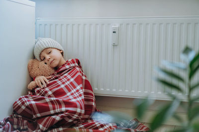 Cute little boy wrapped id plaid wearing knit hat sitting by heater hugging teddy bear, cold at home