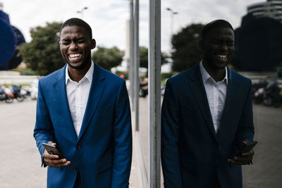 Cheerful male professional holding smart phone while standing by window reflection in city