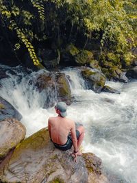 Full length of shirtless man standing on rocks against waterfall