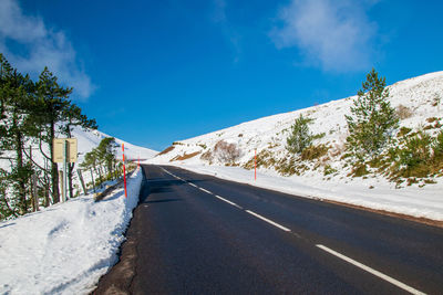 Empty road by snow covered mountains against blue sky