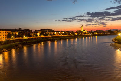 River by illuminated buildings against sky during sunset