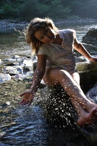 Full length of girl splashing water while sitting on rock by stream