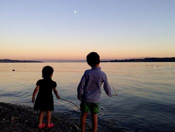 Siblings fishing at riverbank against sky during sunset