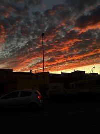 Cars on road by silhouette buildings against sky during sunset