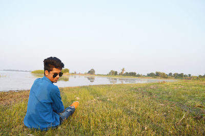 Rear view of man wearing sunglasses sitting on grassy field by lake against sky