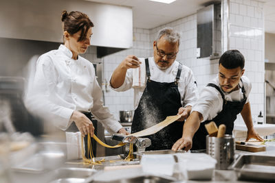 Mature male chef sprinkling flour on dough while using pasta maker with help of female coworker in kitchen at restaurant