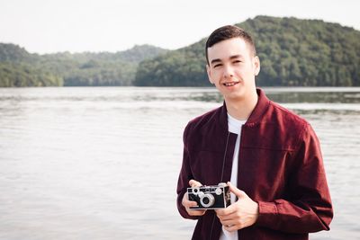 Portrait of young man holding camera against lake and sky