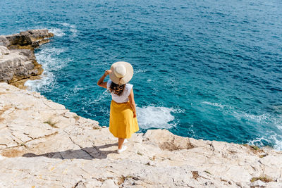 Young woman in summer clothes and hat standing on edge of cliff above sea.