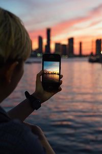 Cropped image of boy photographing city skyline through smart phone during sunset