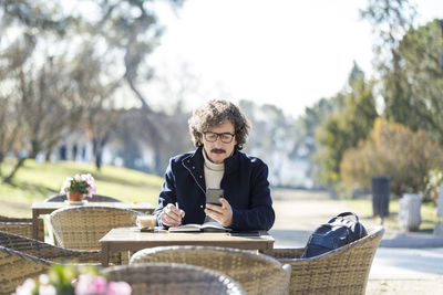 Man using smart phone writing in diary at sidewalk cafe on sunny day