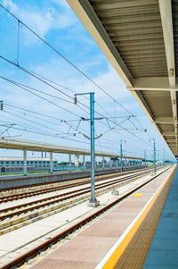 Electricity pylons by railroad station platform against sky during sunny day