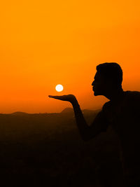 Optical illusion of silhouette man blowing sun against orange sky at sunset