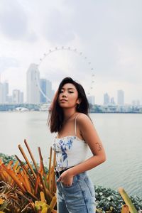 Beautiful young woman standing by river in city against sky