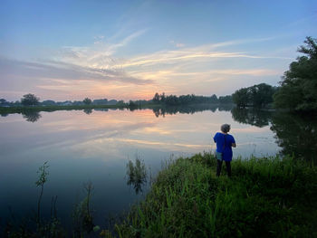 Rear view of man standing by lake against sky during sunset