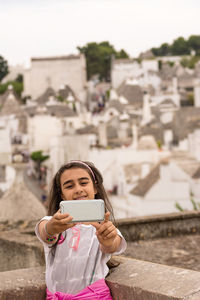 Smiling girl taking selfie while standing against houses