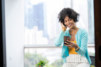 Smiling young woman using mobile phone in office against window