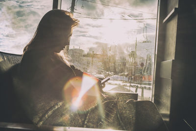 Woman using phone while sitting on sofa by glass window during sunny day