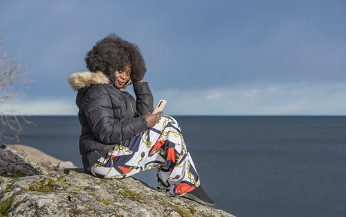 Rear view of woman sitting on rock by sea against sky