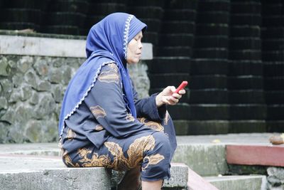 Young woman using mobile phone while sitting on retaining wall