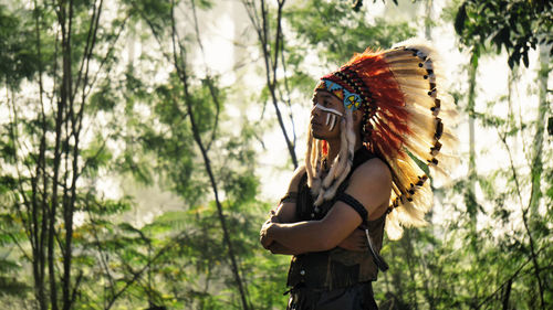 Man wearing feather headdress while standing against trees in forest