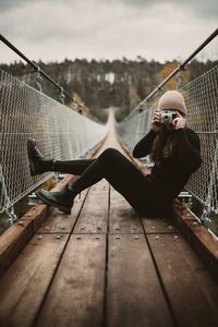 Young woman photographing while sitting on footbridge