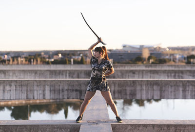 Young female sportsperson practicing sword on structure against clear sky at sunset