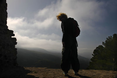 Rear view of hiker standing on cliff by mountains against sky