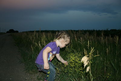 Side view of girl touching flowers at roadside against sky