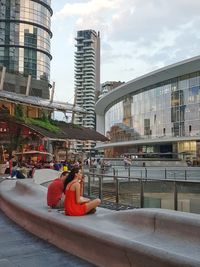 People sitting by modern buildings against sky in city