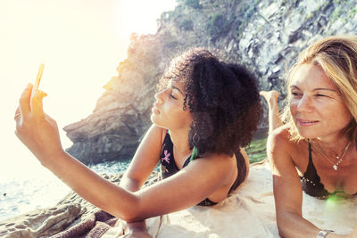 Smiling two women taking selfie while lying on rock formation at sea