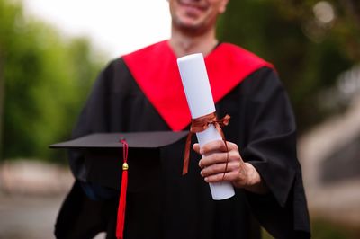Midsection of student wearing graduation gown while holding rolled degree