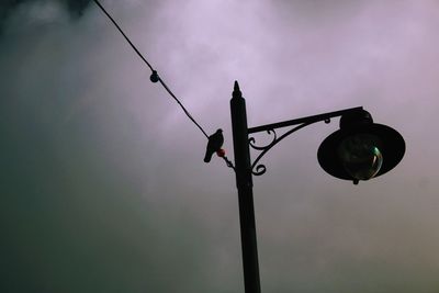 Low angle view of silhouette bird perching on cable against cloudy sky