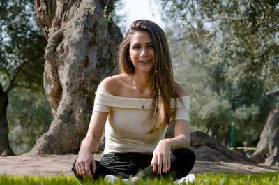 Full length portrait of smiling young woman sitting against trees in park