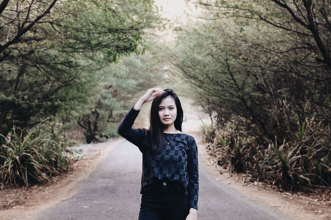 YOUNG WOMAN STANDING ON ROAD AMIDST TREES IN FOREST