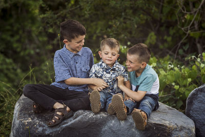 Boys playing with brother while sitting on rock against plants