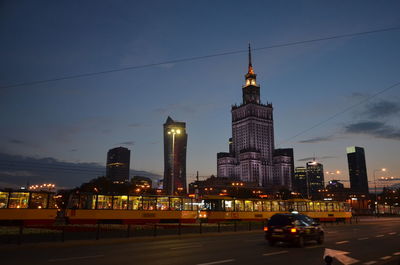 Illuminated buildings by street against sky at night