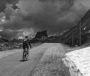 Rear view of man cycling on road by mountains against cloudy sky