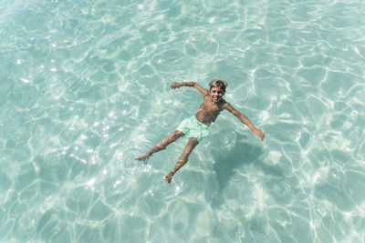 From above of delighted teenage boy swimming in clear sea water on sunny day and looking at camera while enjoying summer vacation