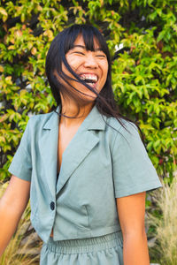 Cheerful young ethnic female in stylish clothes laughing with closed eyes against lush bush on sunny summer day in park