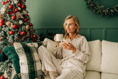 Young woman in the living room uses a mobile phone and drinks coffee on christmas morning.
