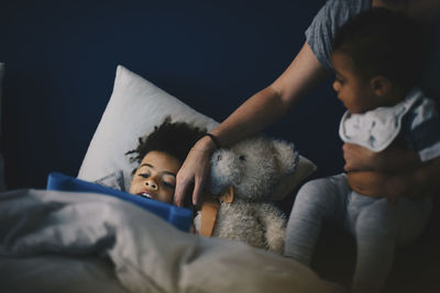 Mother carrying toddler sitting besides son using digital tablet on bed at home