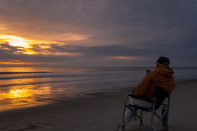 Full length of man sitting on chair at beach against cloudy sky during sunset