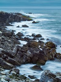 Scenic view of waves splashing on rocks at beach against sky