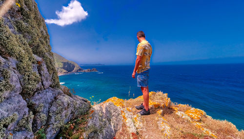 Rear view of man standing on rock by sea against clear blue sky