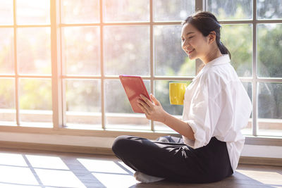 Side view of young woman using phone while sitting on window